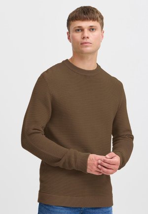 Ung mann med kort hår iført en brun ribbestrikket genser og blå jeans, stående med hendene lett foldet foran en ensfarget lys bakgrunn.