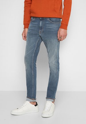 Homme portant un jean bleu délavé avec les ourlets retroussés, des baskets blanches et un sweat-shirt orange, debout devant un fond uni.