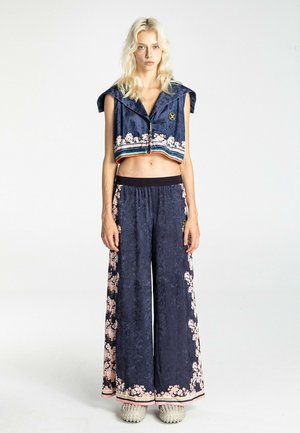 Mujer rubia con un top corto azul marino sin mangas y pantalones de pierna ancha con estampado floral rosa, de pie sobre un fondo blanco.