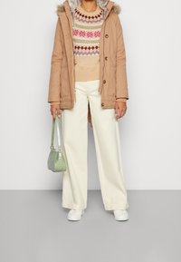 Béžová parka s kožešinovým lemem, vzorovaný svetr v růžové a zelené, krémové kalhoty s širokými nohavicemi a malá zelená kabelka. Celý vzhled doplňují bílé tenisky.