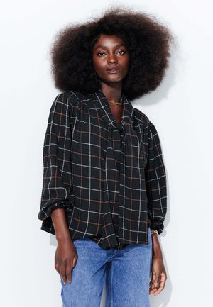 Blouse à carreaux noirs avec des lignes blanches et orange, dotée d'un col noué et de manches bouffantes, assortie à un jean bleu. Tissu texturé.