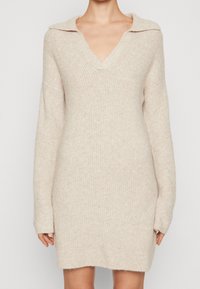 Stickad klänning i ljusbeige med V-ringning och krage, med långa ärmar och lös passform, mjuk textur och ribbat mönster.