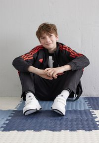 Musta urheilupuku punaisilla yksityiskohdilla, valkoiset kengät, istuu kuvioiduilla sinivalkoisilla laattoilla. Taustalla vaaleanharmaa seinä.