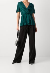 Blusa avvolgente a maniche corte color teal con orlo a peplum, abbinata a pantaloni neri a gamba larga. Il modello tiene in mano una piccola borsa nera.