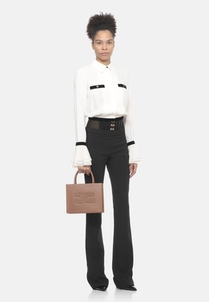 Donna in piedi che indossa una camicetta bianca con dettagli neri, pantaloni neri a vita alta, scarpe nere con punta affilata, che tiene una borsa marrone.