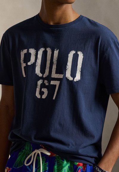 Άνδρας που φοράει μπλε ναυτικό t-shirt με φθαρμένο κείμενο "POLO 67" και τροπικά φλοράλ σορτς με λευκό κορδόνι, στέκεται με το ένα χέρι στην τσέπη.