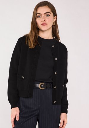 Femme aux cheveux bruns portant des boucles d'oreilles creoles dorées, un cardigan noir boutonné, une chemise noire et un pantalon bleu marine à rayures fines avec une boucle de ceinture décorative.