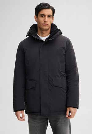 Young man wearing a black hooded winter jacket with flap pockets and a zipper, standing against a plain light background.