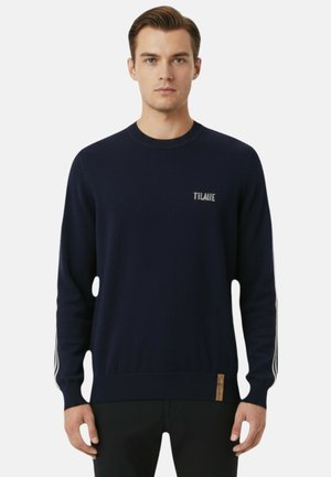 Uomo che indossa un maglione girocollo blu navy con la scritta "TILAJIE" sul petto e strisce bianche sulle maniche, in piedi davanti a uno sfondo neutro.