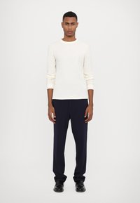 Lys beige ribbet langermet skjorte kombinert med mørk marinebukser. Outfiten har rund hals og tilpassede mansjetter, med svarte sko.