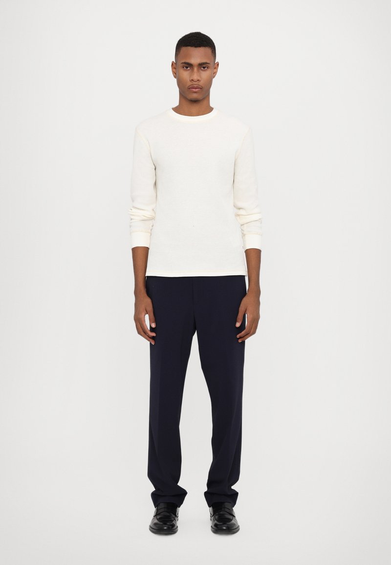 Lys beige ribbet langermet skjorte kombinert med mørk marinebukser. Outfiten har rund hals og tilpassede mansjetter, med svarte sko.