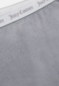 Detailný záber na svetlo šedú textúrovanú látku s bielym elastickým pásom, na ktorom je embosovaný nápis "Juicy Couture" v gotickom písmovom štýle.