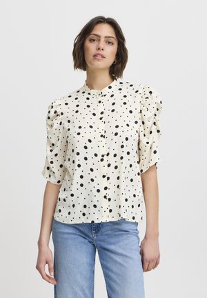 Cremefarvet bluse med sorte prikker, korte puffærmer og knaplukning foran. Blødt stof med en tekstureret overflade. Kombineret med lyseblå jeans.