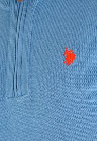 Svijetloplavi pletivi polo majica sa kratkim zipom i izvezenim narančastim logom jahača i konja na lijevoj strani grudi.