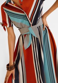 Robe rayée avec devant boutonné, manches courtes à nouer, taille ceinturée, et couleurs comprenant rouge, sarcelle, marine et gris.