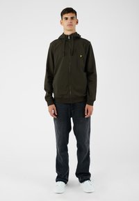 Giacca a zip verde scuro con cappuccio, con un piccolo logo giallo sul petto. Abbinata a jeans scuri e sneaker bianche.