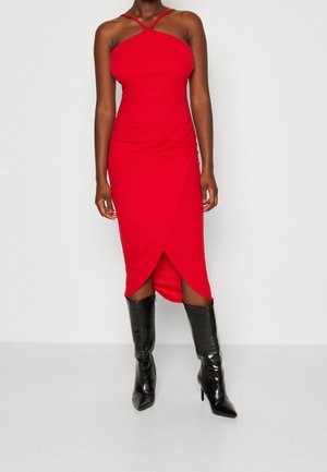 Femme portant une robe midi rouge à dos nu avec un ourlet tulipe, associée à des bottes noires brillantes pointues hauteur genou.