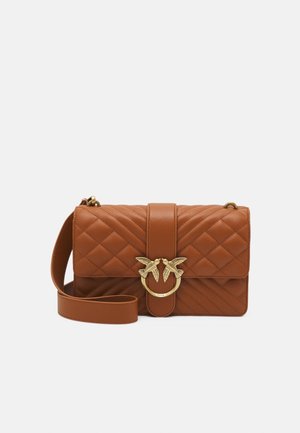 Hnědá prošívaná kožená crossbody taška se zlatým dvojitým kroužkem ve tvaru ptáka a nastavitelným popruhem na jednobarevném bílém pozadí.