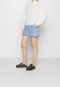 Blusa bianca in pizzo con maniche a sbuffo, abbinata a pantaloni corti in denim azzurro chiaro con tasche frontali e cuciture decorative, e scarpe nere slip-on.