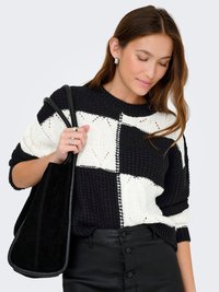 Pulover tricotat alb-negru cu un model în blocuri, purtat cu pantaloni negri. O geantă neagră, lejeră, este ținută de model.