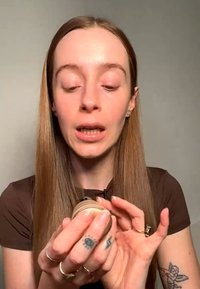 Young woman with long hair holding and examining a small jar, speaking or explaining, wearing rings and visible tattoos on hands and arm.
