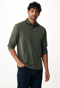 Olijfgroene polo met lange mouwen, voorzien van een klassieke kraag en een subtiel logo, gecombineerd met donkere spijkerbroek. Zacht materiaal, relaxed fit.