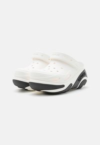 White clogs with perforations, featuring a black rubber sole and heel accent. Slip-on design with a back strap for secure fit.
