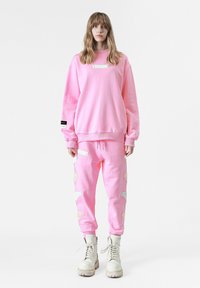 Sudadera oversized de color rosa claro con puños acanalados, combinada con joggers a juego que presentan patrones de alas texturizados. Botas de combate blancas completan el conjunto.