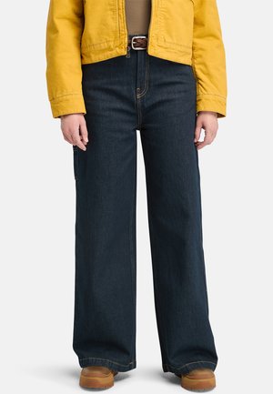Pantalons en denim foncé à jambes larges avec des coutures visibles, complétés par une veste jaune courte. Une ceinture marron et des chaussures beige achèvent le look.