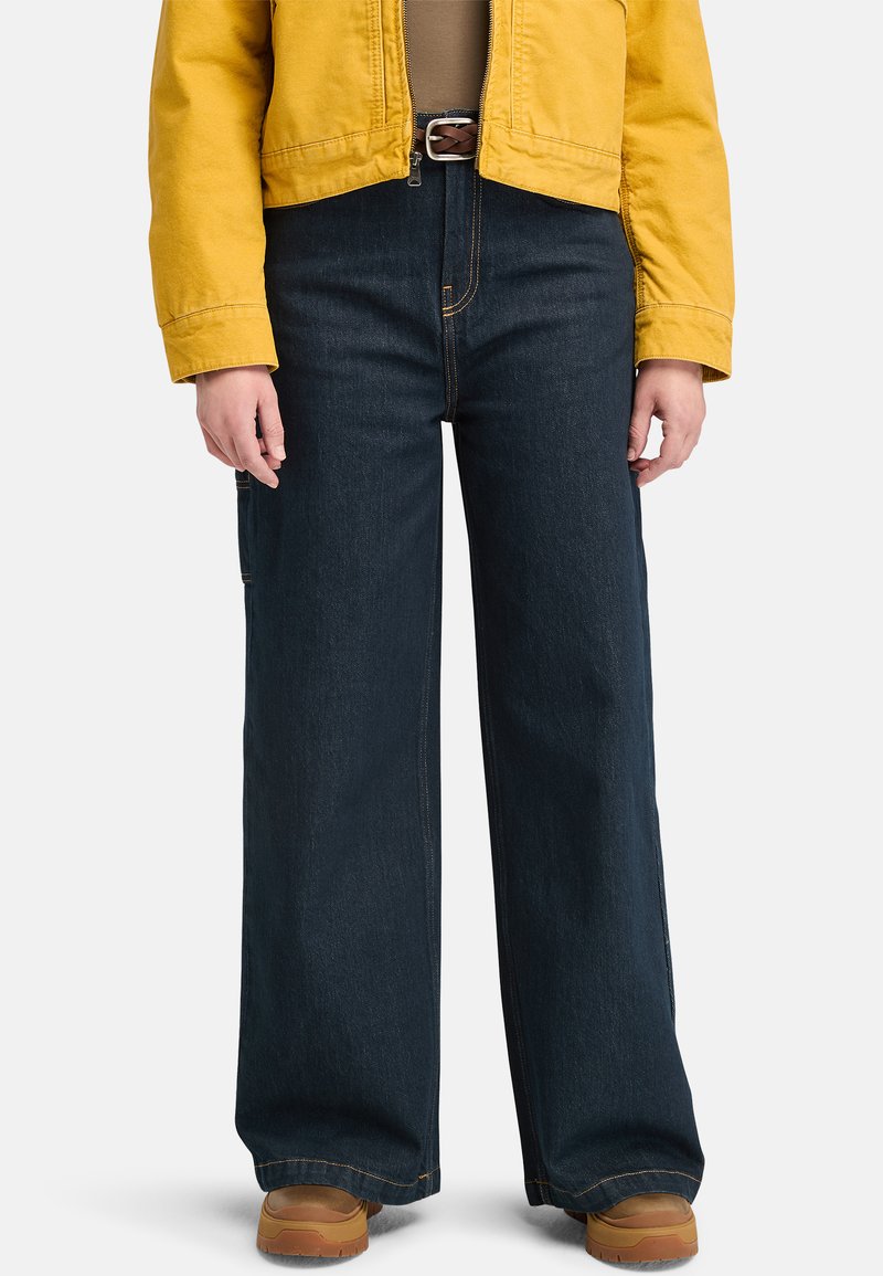 Pantalons en denim foncé à jambes larges avec des coutures visibles, complétés par une veste jaune courte. Une ceinture marron et des chaussures beige achèvent le look.