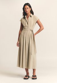 Bež midi haljina s ovratnikom, kratkim kap rukavima, remenom za vezanje i plisiranom suknjom. Stilizirana s crnim ravnim sandalama.