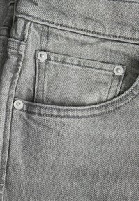 Szary denim z przednią kieszenią z srebrnym metalowym elementem i widocznym przeszyciem. Teksturowana powierzchnia o klasycznym wzorze dżinsów.