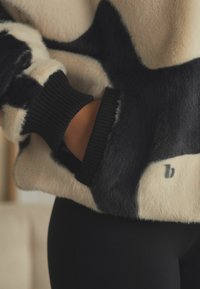 Kunstlikust karusnahast pullover kreemja ja musta värviga, millel on sooniline must mansett ja diskreetne logo detail. Pehme tekstuur ja stiilne muster.