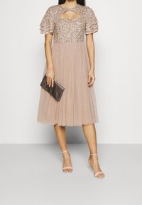 Vestido de noche beige con lentejuelas, mangas cortas con volantes, escote en forma de ojal y falda de tul plisada. Combinado con un clutch oscuro y tacones nude.