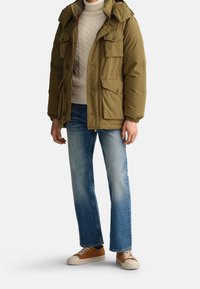 Olivengrøn pufferjakke med flere forlommer, ribstrikkede manchetter og aftagelig hætte, båret over en cremefarvet kabelstrikket sweater og blå jeans.