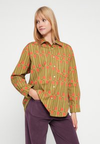 Πράσινο μπλουζάκι με κόκκινα λουλουδένια σχέδια, κλείσιμο με κουμπιά, μία τσέπη, μακριά μανίκια, άνετη εφαρμογή, συνδυασμένο με μοβ παντελόνι.