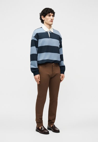 Νεαρός άνδρας στέκεται φορώντας μπλε και ναυτικό ριγέ μακρυμάνικο πόλο, καφέ στενά παντελόνια και σκούρα καφέ μοκασίνια μπροστά σε απλό φόντο.