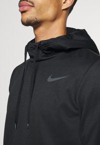 Gros plan sur une personne portant un sweat à capuche noir Nike avec cordons, montrant une vue partielle du visage et le logo Nike sur la poitrine.