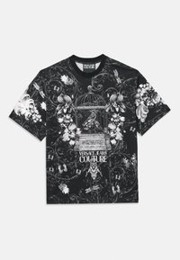 Čierne tričko s krátkym rukávom, na ktorom sú zložité biele vzory kvetov a vtáčich klietok, s textom "VERSACE JEANS COUTURE" na spodnej strane.