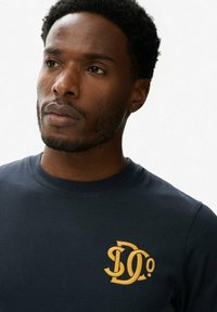 Marineblåt langærmet t-shirt lavet af blød bomuld, med et gult logo på venstre bryst. Rund halsudskæring med en tætsiddende pasform.