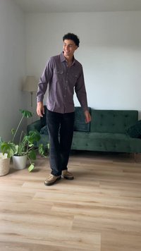 Μοντέλο που φοράει ένα μωβ πουκάμισο με κουμπιά και μαύρο παντελόνι, στέκεται σε ξύλινο πάτωμα δίπλα σε μια πράσινη καναπέ και φυτά σε γλάστρες.