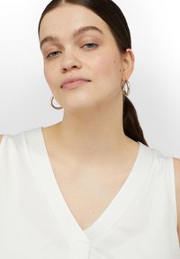 Biała, bezrękawnikowa bluzka z dekoltem w kształcie litery V; gładki materiał o lekkim połysku. Srebrne kolczyki-wkrętki uzupełniają całość. Prosta i elegancka.