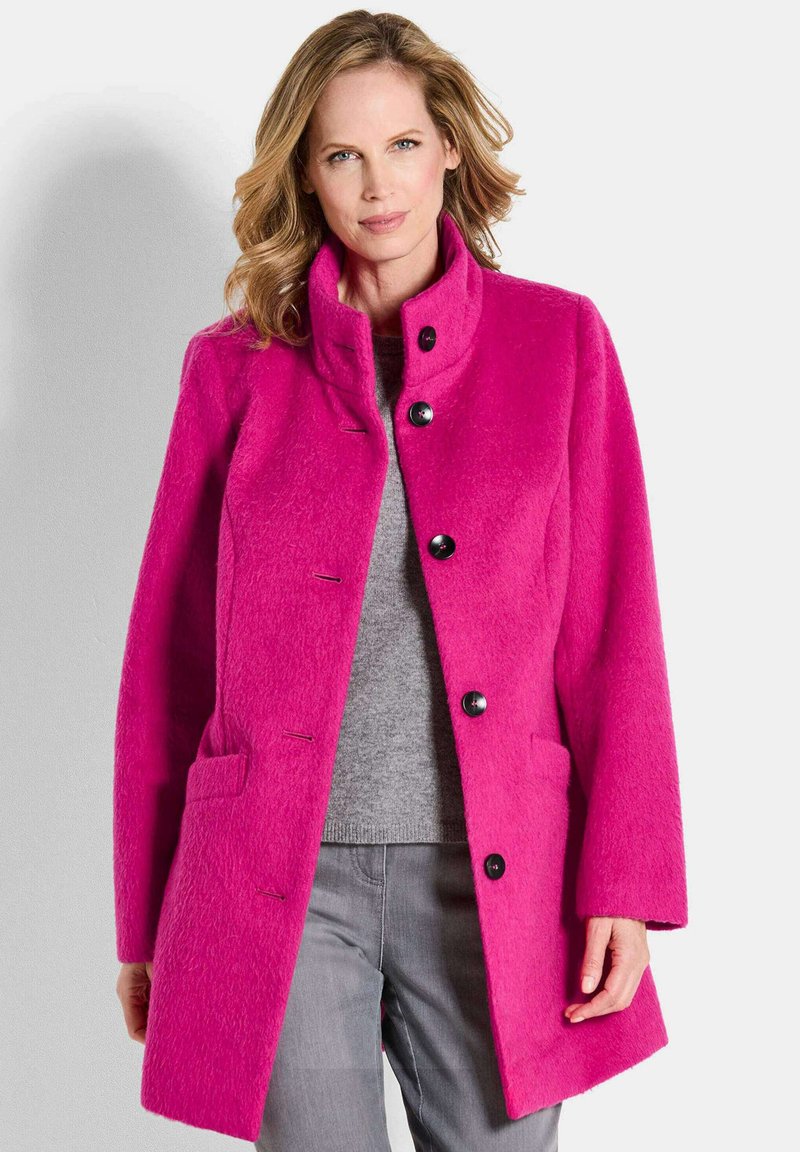 Cappotto di lana fucsia con colletto alto, dotato di bottoni neri, tasche laterali e una finitura testurizzata. Indossato sopra un maglione grigio.