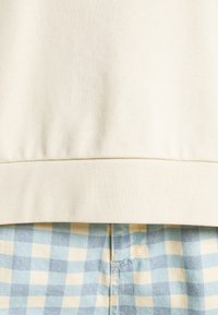 Il bordo di una felpa in cotone crema si trova sopra dei pantaloni corti a scacchi blu e beige, caratterizzati da una texture morbida e un elastico in vita.