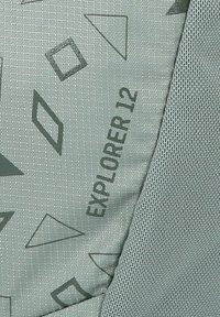 Tela de mochila verde clara con patrones geométricos. Texto "EXPLORER 12" impreso en verde oscuro. Acento de malla texturizada en un lado.