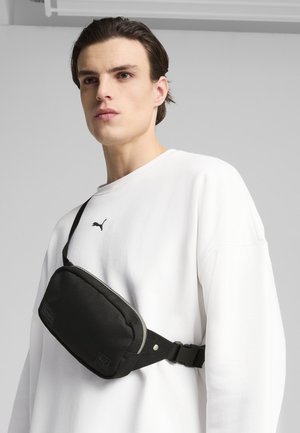 Zwarte stoffen heuptas met een ritscompartiment, zilveren afwerking, verstelbare riem en een logopatch. Draagt over een wit sweatshirt.