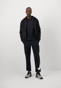 Sudadera negra con cremallera y detalles texturizados, combinada con una camiseta negra de manga corta y pantalones negros con un logo blanco de Nike. Zapatillas gris.