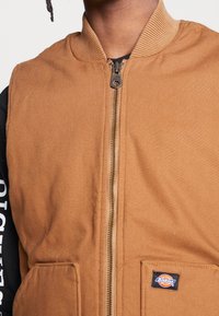 Brązowa kamizelka z płótna z prążkowanym kołnierzem, zamkiem błyskawicznym z przodu, dwoma kieszeniami i małą metką w prawym dolnym rogu.