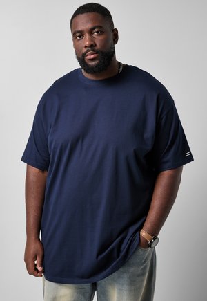 Uomo nero con barba che indossa una maglietta blu navy e jeans chiari, in piedi con una mano in tasca contro uno sfondo chiaro e uniforme.