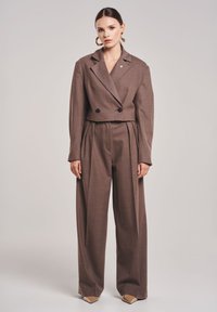 Bruin op maat gemaakte cropped blazer met dubbele zwarte knopen, gecombineerd met losse bruine broek. Soepele textuur en gestructureerd ontwerp. Chunky gouden oorbellen.